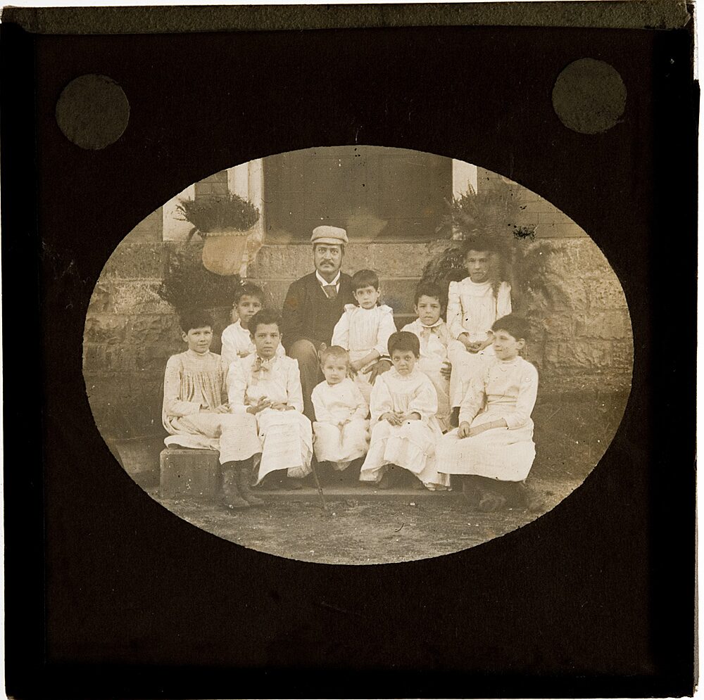 A lantern slide in an oval frame: a man sits on a stone step with eight children in white clothes.